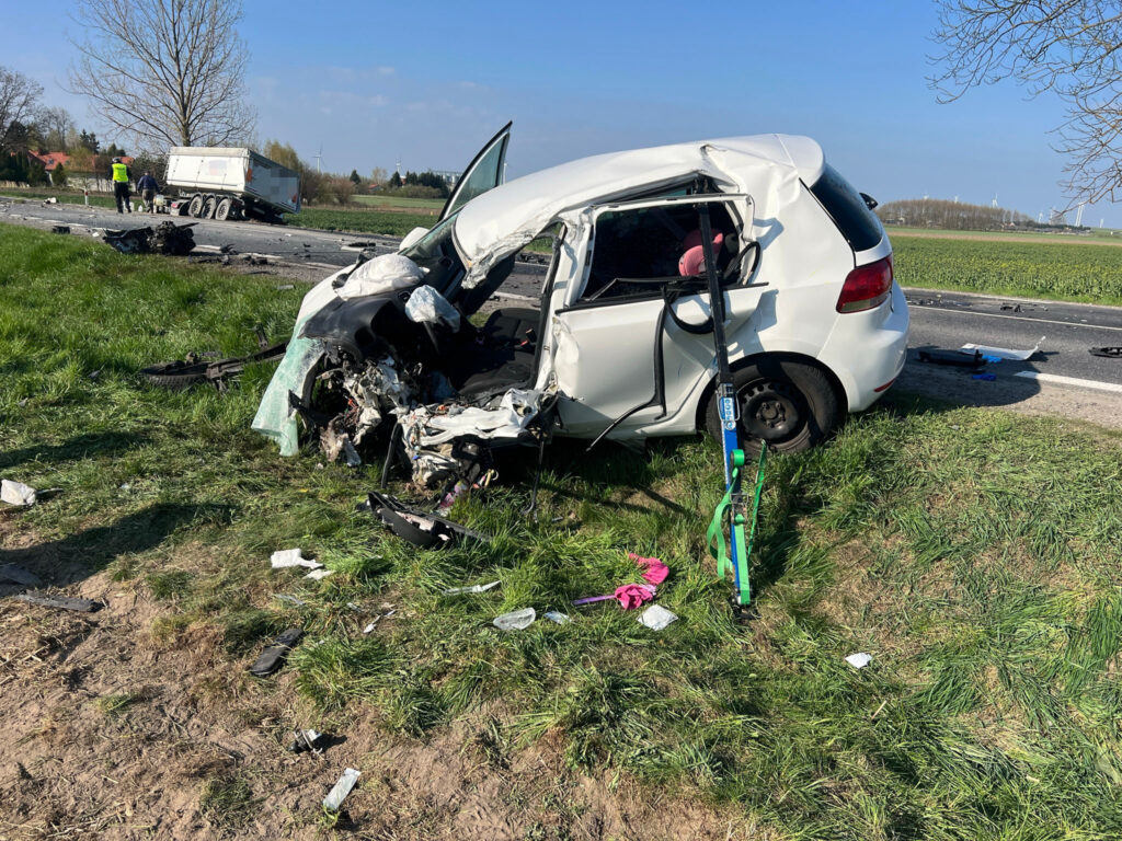 Zniszczony samochód osobowy leży na poboczu drogi, obok pracujący zespół ratunkowy oraz porozrzucane fragmenty wraku.