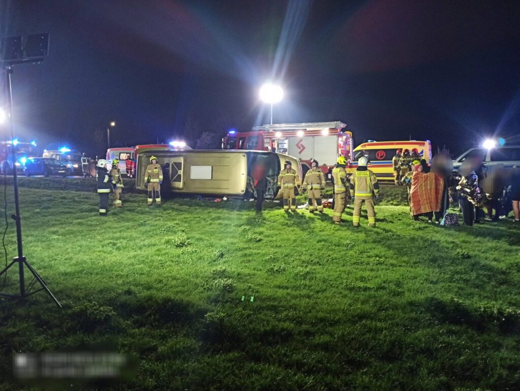 Grupa strażaków udziela pomocy przy przewróconym pojeździe na trawie w nocy, otoczona przez pojazdy ratunkowe.