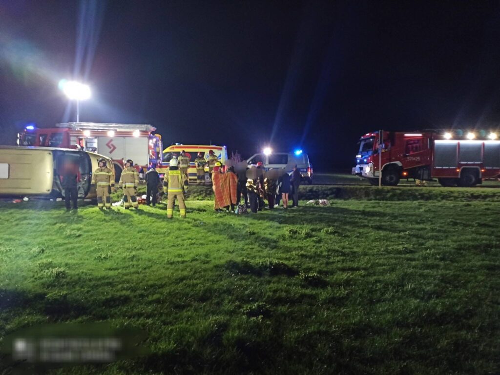 Grupa ratowników i strażaków udziela pomocy poszkodowanym przy przewróconym samochodzie na trawie w nocy.