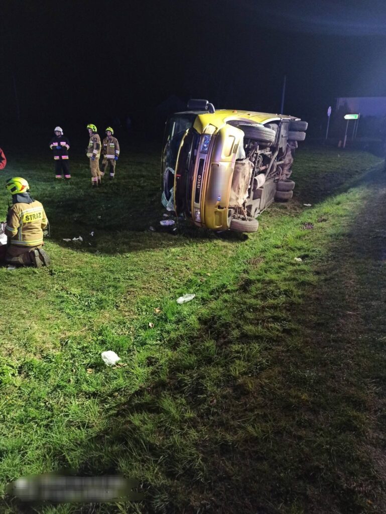 Żółty pojazd dostawczy leży na boku na trawie, a obok niego stoją strażacy w mundurach, prowadząc działania ratunkowe.