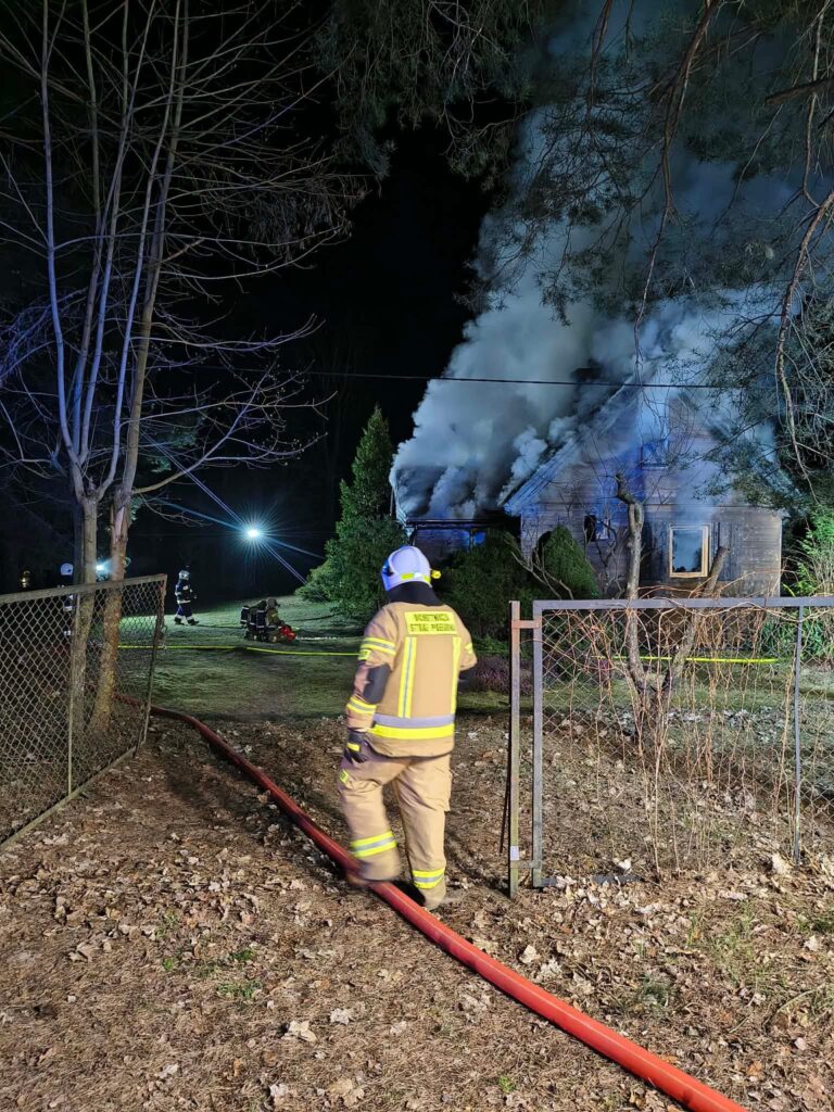 Strażak w mundurze z wężem gaśniczym podchodzi do palącego się budynku w nocy, w tle widoczne płomienie i dym.