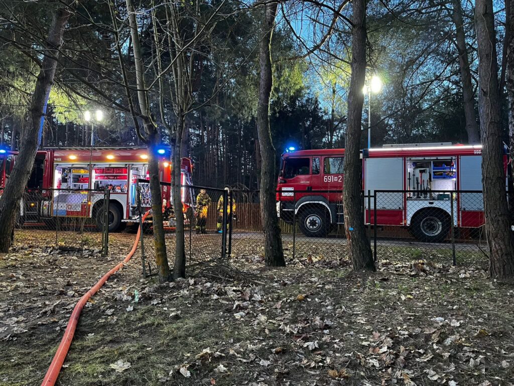 Strażacy pracują przy wozie strażackim w lesie, otoczeni drzewami i sprzętem gaśniczym.