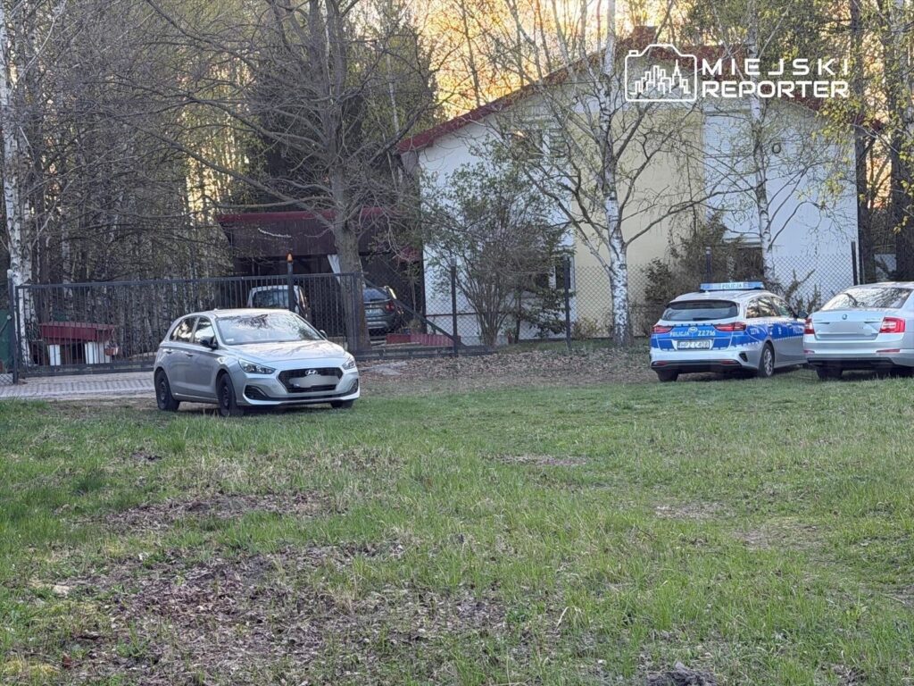Na trawiastej przestrzeni stoi srebrny samochód osobowy, obok zaparkowane dwa radiowozy policyjne przy ogrodzeniu. W tle widoczny jest dom otoczony drzewami.