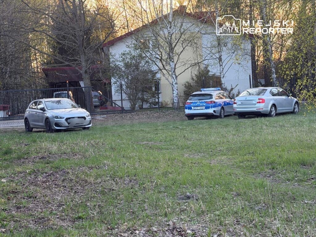 Na trawie parkuje srebrny samochód osobowy obok dwóch radiowozów policyjnych, w tle widać dom.