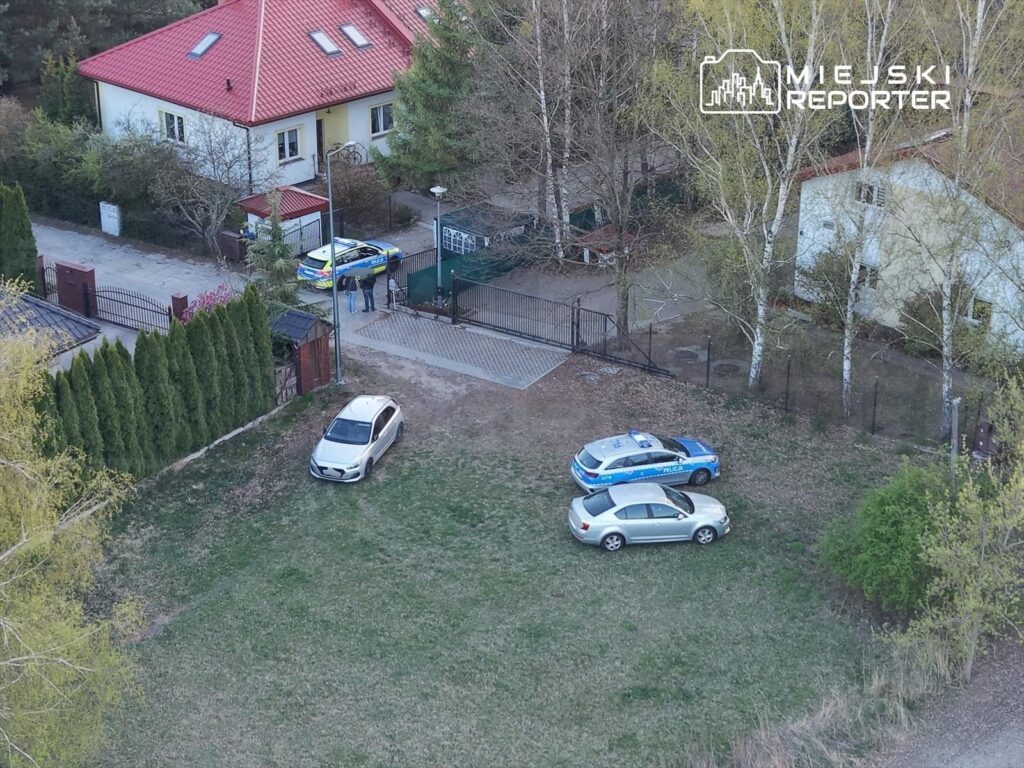 Dwa samochody policyjne stoją przed ogrodzeniem, obok rozmawiający mężczyzna i kobieta, w tle dom z czerwoną dachówką.