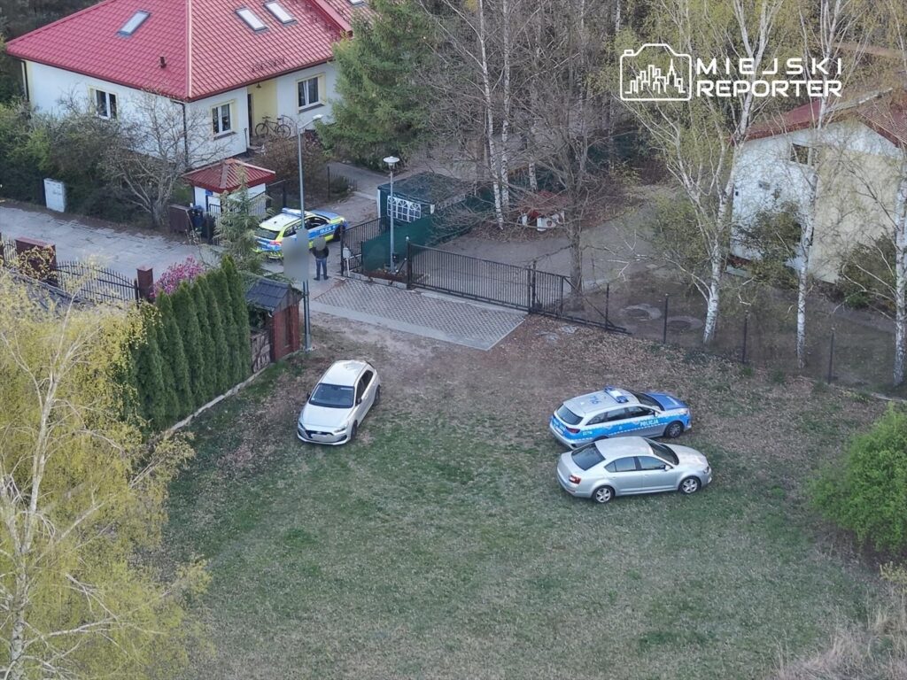 Mężczyzna w mundurze rozmawia z policjantem obok radiowozu przed domem z czerwonym dachem.