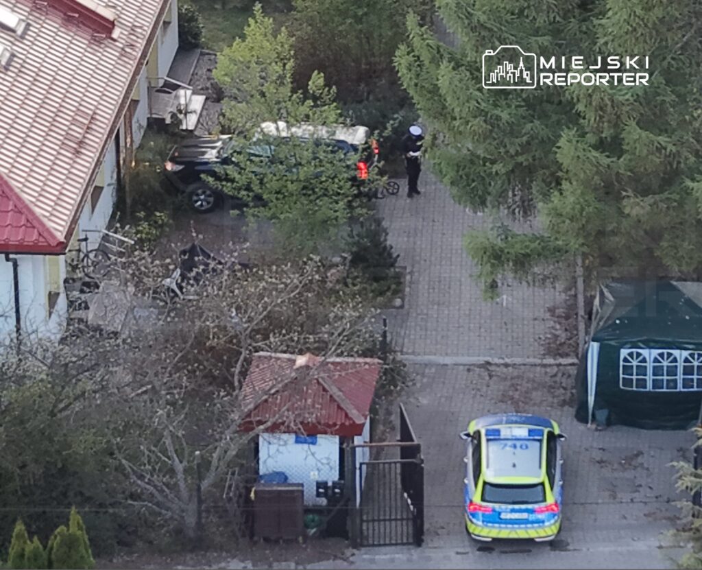 Policjant rozmawia z kierowcą samochodu zaparkowanego na podjeździe przed domem. W tle widoczny zielony namiot.