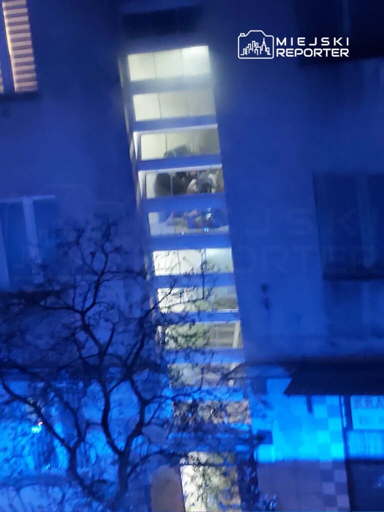 Grupa ludzi w jasnym pomieszczeniu z dużymi oknami, oświetlonym niebieskim światłem, siedzi przy stole.