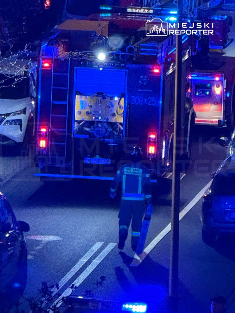 Strażak w mundurze przechodzi obok wozu strażackiego na ulicy oświetlonej niebieskimi światłami.