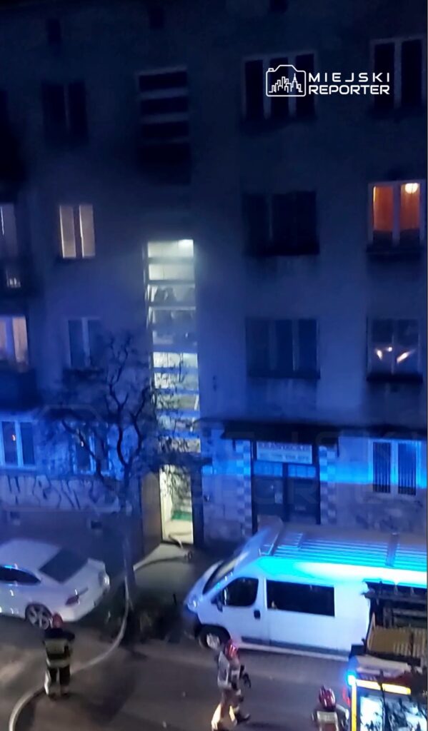 Strażacy w hełmach gaszą pożar w budynku mieszkalnym, obok zaparkowane białe auto i niebieski wóz strażacki.