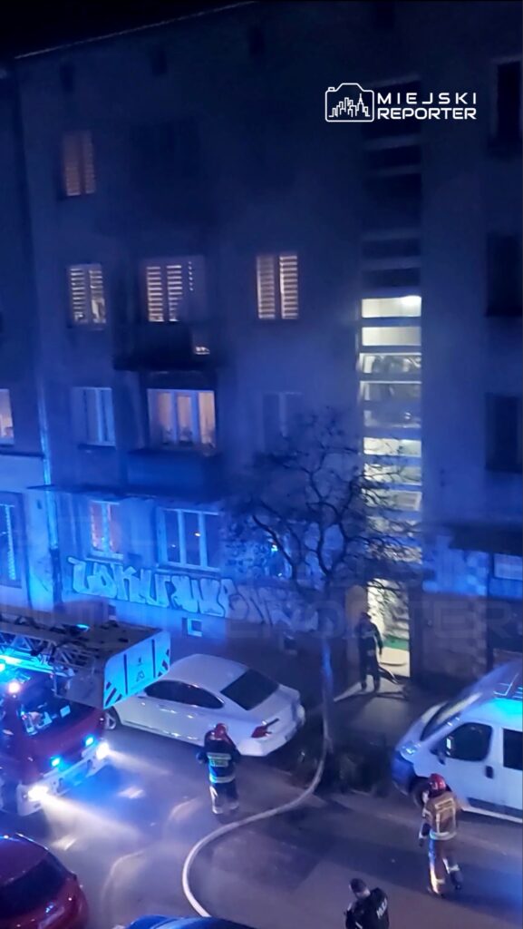 Strażacy w hełmach oraz mundurach, pracujący przy wozach strażackich, gaszą pożar w budynku mieszkalnym w nocy.