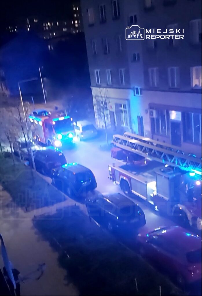 W nocy na ulicy stoją dwa wozy strażackie z włączonymi sygnałami świetlnymi, otoczone przez zaparkowane samochody.