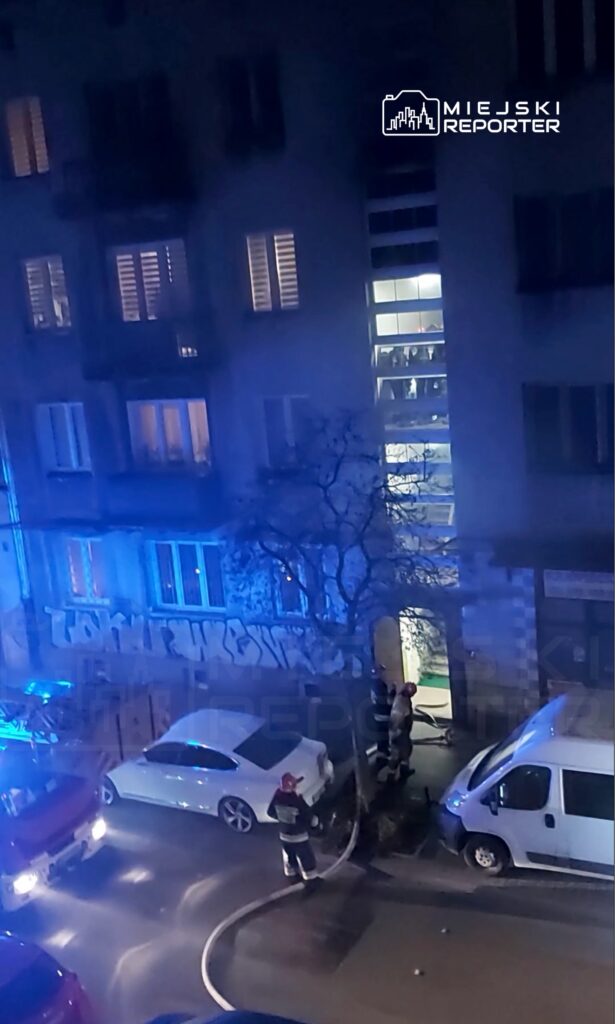 Grupa strażaków z wężem gaśniczym w pobliżu zaparkowanych pojazdów na ulicy, w tle oświetlone okna budynku.