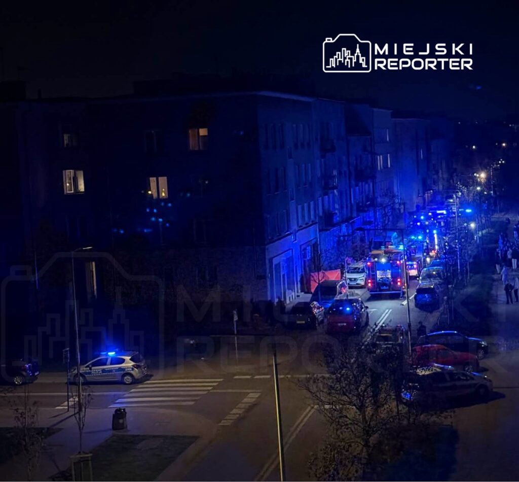 W nocy na ulicy widać pojazdy służb ratunkowych z włączonymi sygnałami świetlnymi oraz zebrane osoby.