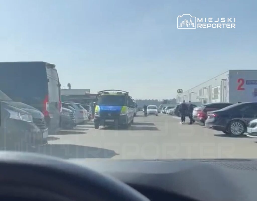 Policyjny radiowóz przejeżdża przez parking, na którym stoją zaparkowane samochody, w tle widać budynki.