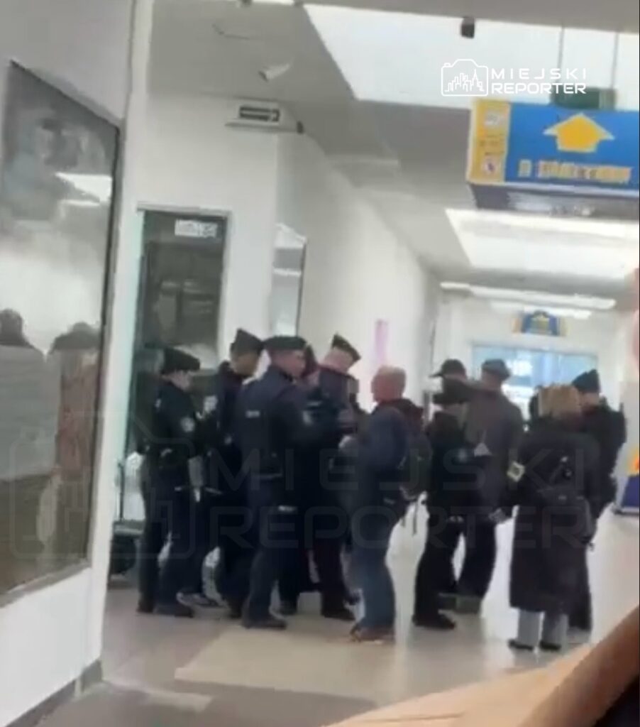 Grupa policjantów rozmawia z mężczyzną w towarzystwie kilku osób w korytarzu budynku publicznego.