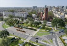 Nowoczesny węzeł komunikacyjny z torowiskiem tramwajowym, zielenią i zabytkowym kościołem w tle.