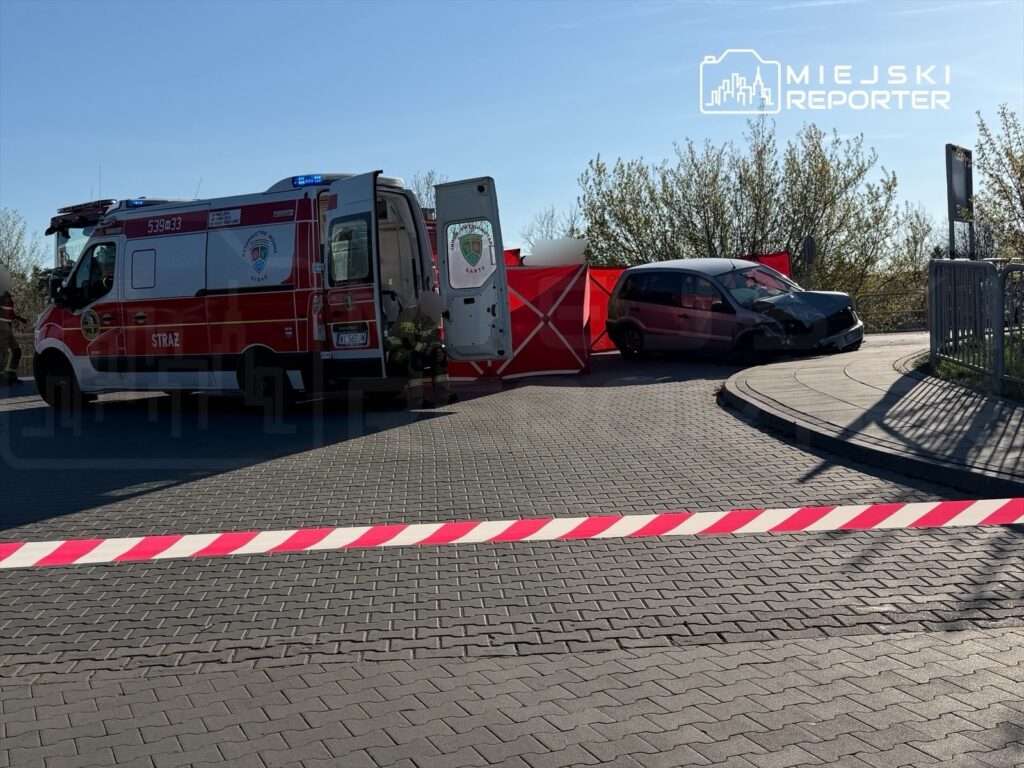 Czerwony ambulans z włączonymi sygnałami stoi obok zderzonego czarnego samochodu, a teren jest zabezpieczony taśmą.
