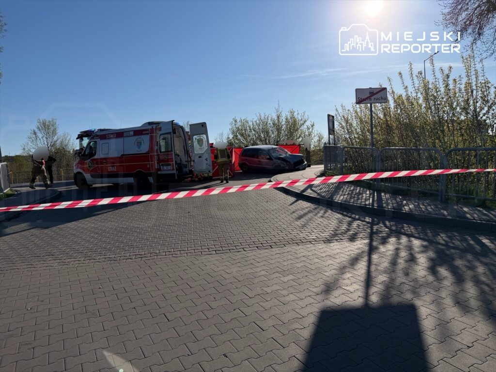 Pojazd osobowy z uszkodzeniami stoi obok karetki, a w tle widoczne jest ogrodzenie z taśmą ostrzegawczą.