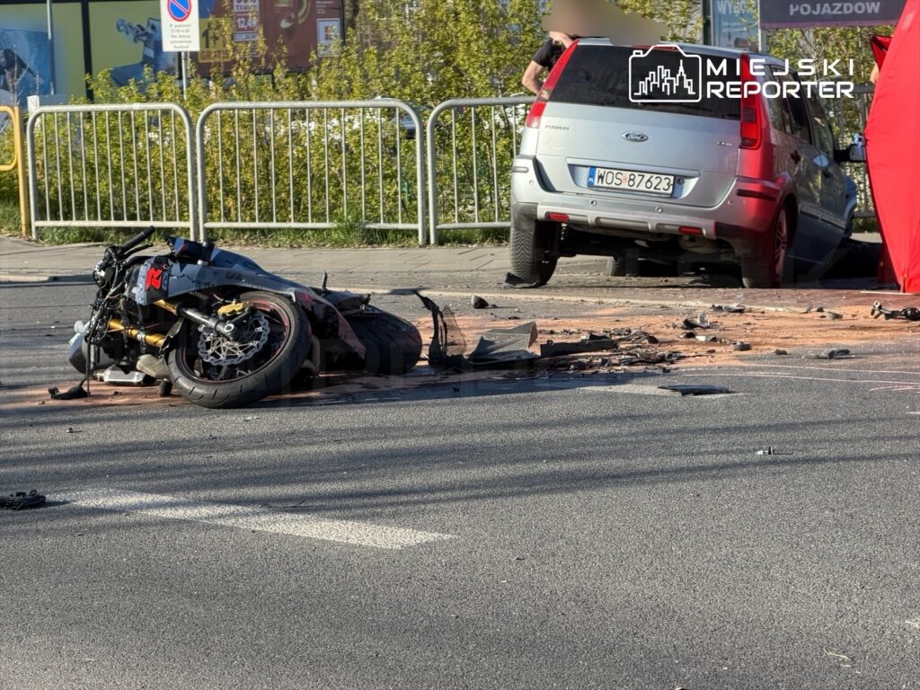 Uszkodzony motocykl leży na jezdni obok zaparkowanego samochodu, wokół widoczne fragmenty rozbitego pojazdu.