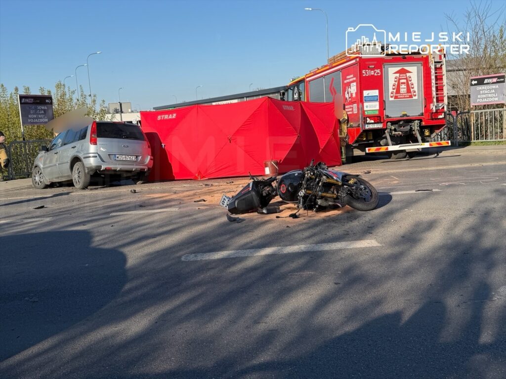 Motocykl leży na jezdni obok uszkodzonego samochodu, a w tle widać wóz strażacki oraz czerwony namiot zabezpieczający miejsce zdarzenia.