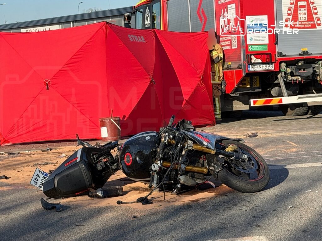 Motocykl leżący na jezdni po wypadku, obok czerwony namiot ratunkowy i wóz strażacki. Na drodze widoczne ślady.