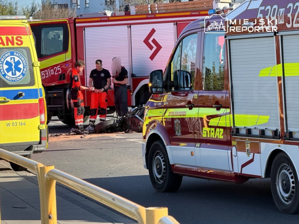 Ratownicy medyczni oraz strażacy pracują nad zabezpieczeniem miejsca wypadku na ulicy, obok dwóch pojazdów służb ratunkowych.