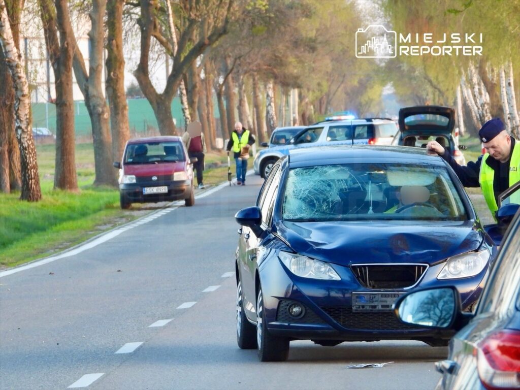 Na drodze stoi niebieski samochód z uszkodzoną szybą, obok pracują policjanci oraz świadkowie zdarzenia.