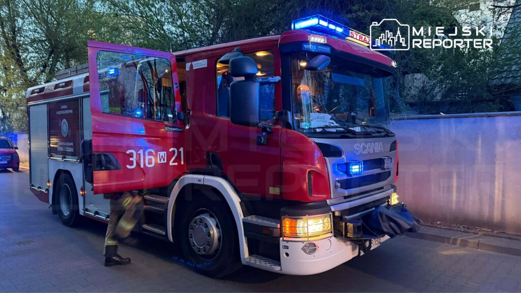 Czerwony wóz strażacki z włączonymi sygnalizatorami świetlnymi stoi na ulicy, obok niego strażak w mundurze.