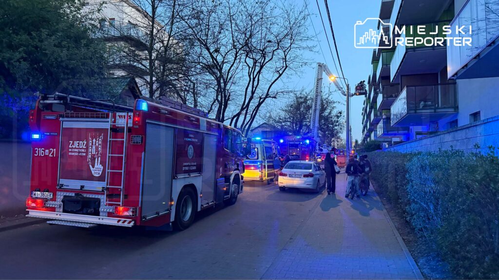 W tle widoczny nowoczesny budynek mieszkalny, a na pierwszym planie pojazdy straży pożarnej z włączonymi sygnałami świetlnymi.