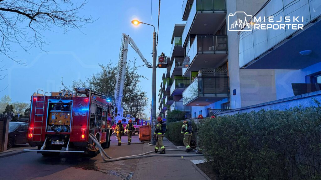 Strażacy w mundurach pracują przy wozie strażackim, rozkładając węże na chodniku obok bloku mieszkalnego.