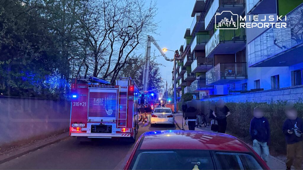 Straż pożarna z włączonymi sygnałami świetlnymi interweniuje przy bloku mieszkalnym, a obok zgromadził się tłum ludzi.