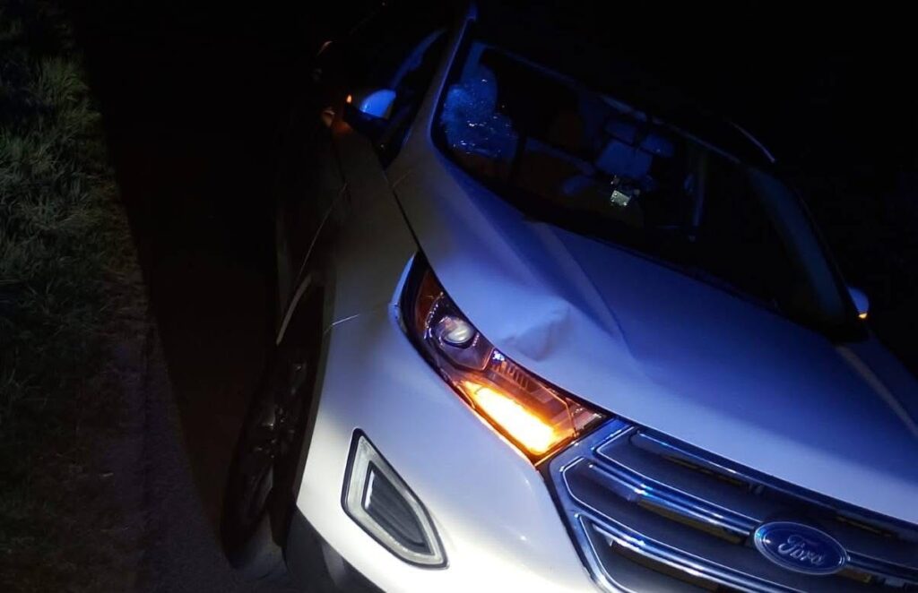 Biały samochód osobowy z uszkodzoną maską stoi na poboczu drogi w nocy, z włączonymi światłami.