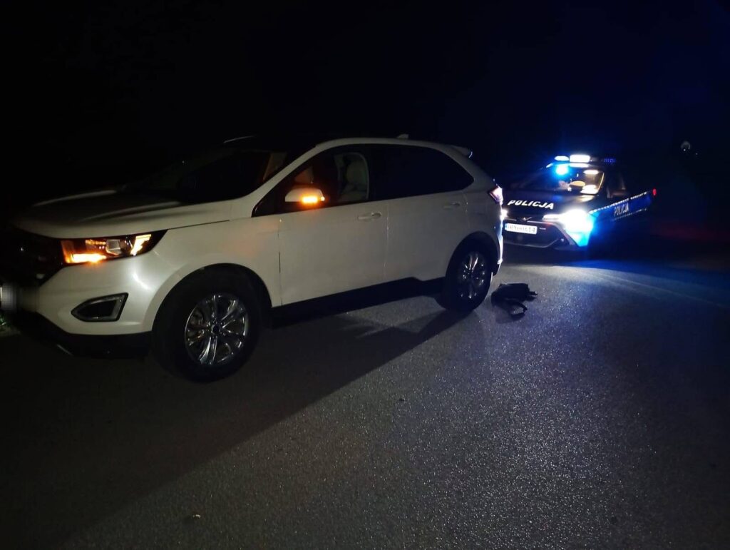 Biały SUV zatrzymany na drodze, obok policyjny radiowóz z włączonymi sygnałami świetlnymi.