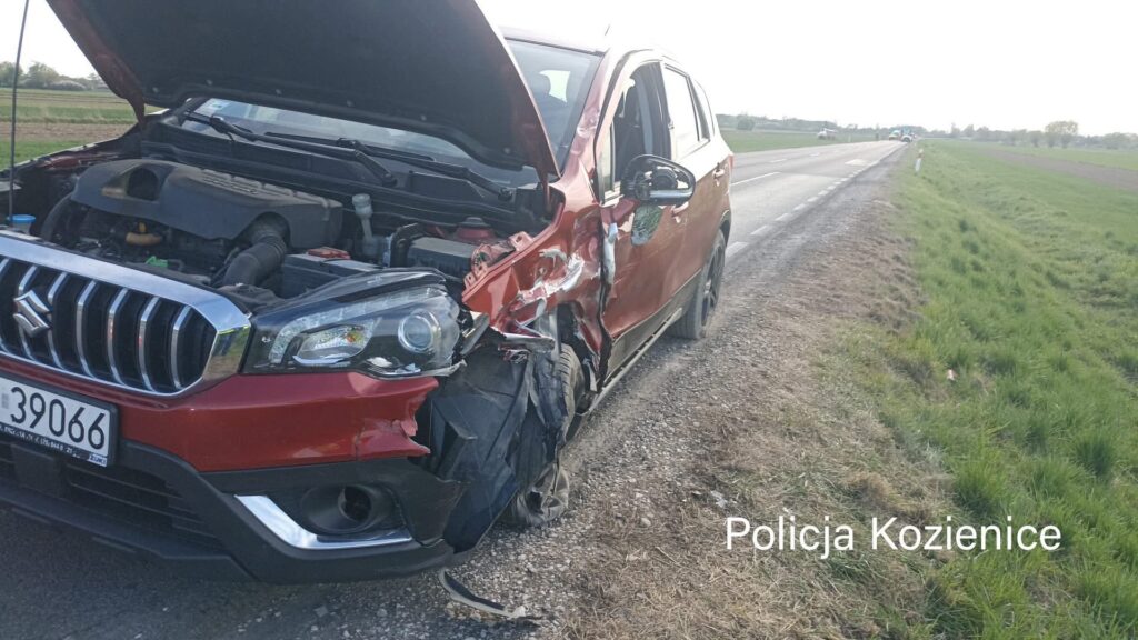 Uszkodzony czerwony samochód osobowy z wgniecioną karoserią stoi przy drodze, obok otwartego silnika.