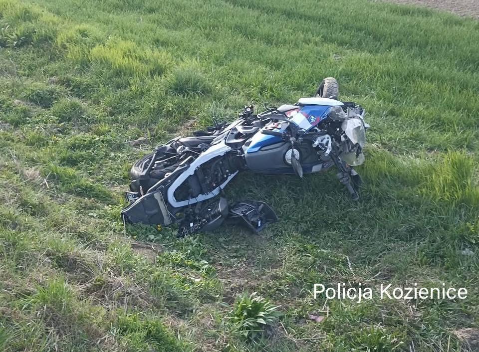 Motocykl leżący na boku na zielonym polu, otoczony trawą i niską roślinnością.