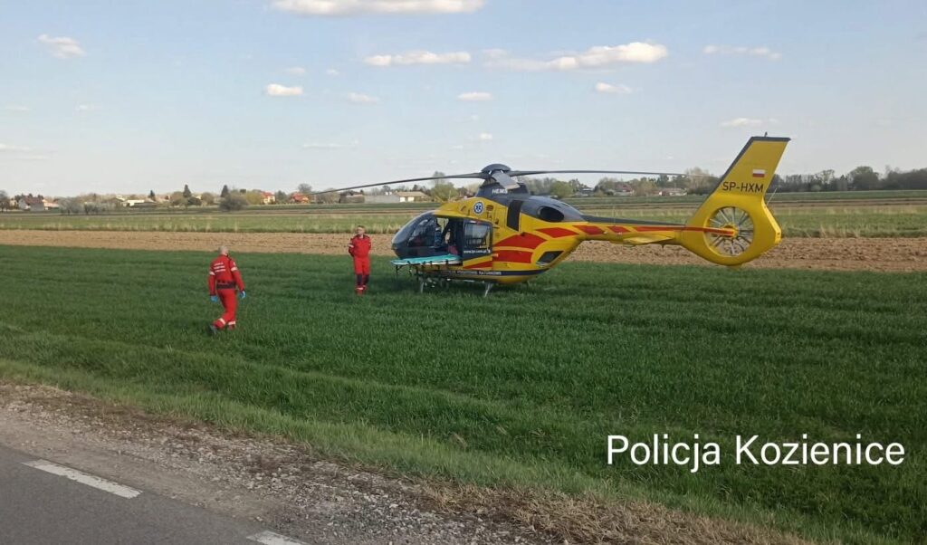 Ratownicy w czerwonych strojach podchodzą do lądowiska helikoptera na zielonym polu.