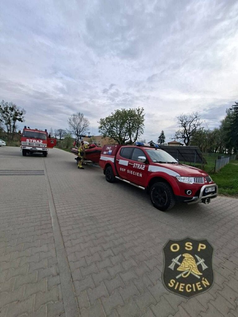 Strażacy z OSP Siecień przygotowują sprzęt ratunkowy obok samochodu strażackiego na drodze w otoczeniu drzew.