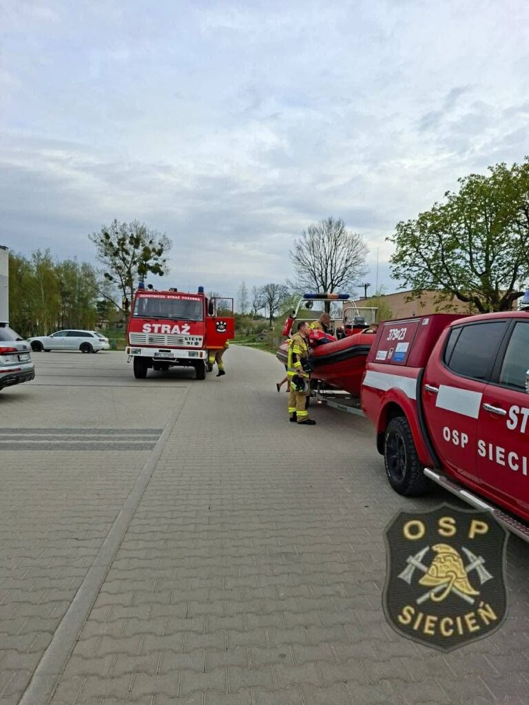 Strażacy w mundurach przygotowują łódź do transportu przy wozach strażackich na parkingu.