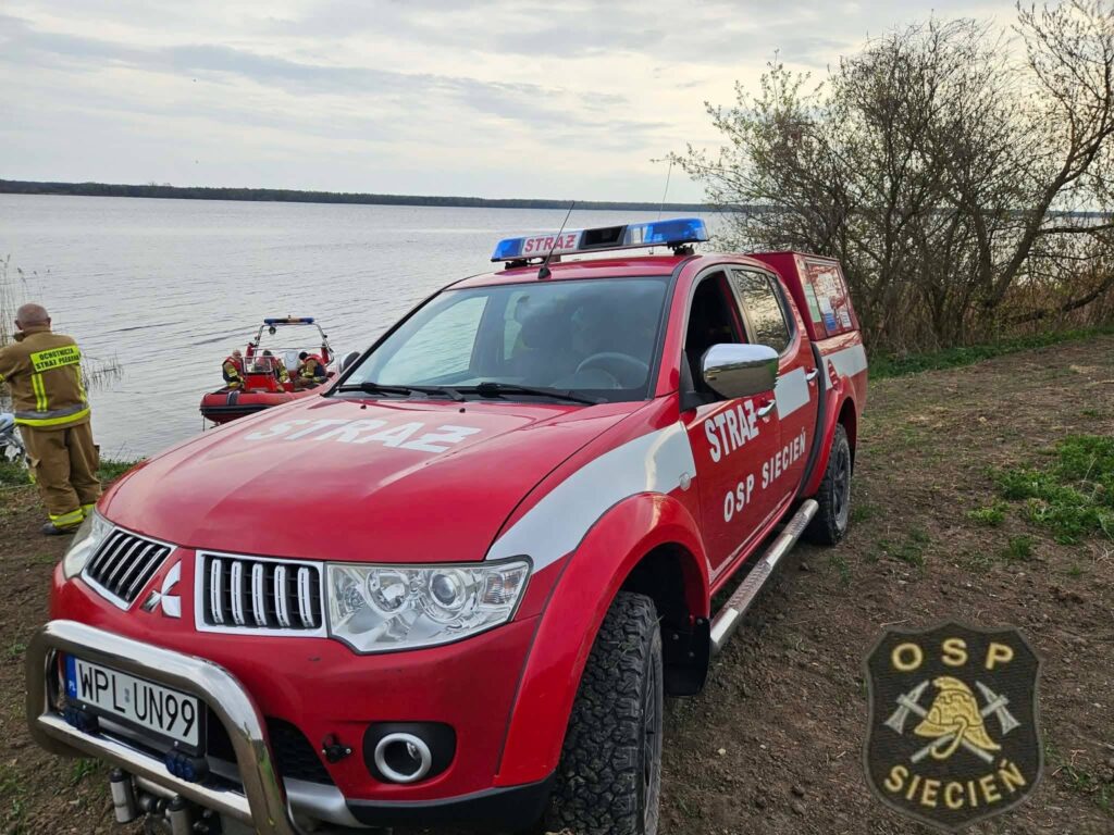 Czerwony samochód strażacki stoi na brzegu jeziora, a w tle widać zespół ratowników w pontonie.