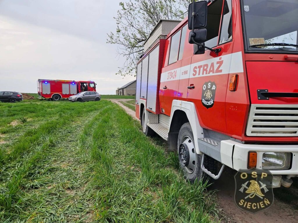 Czerwony wóz strażacki z włączonymi sygnałami świetlnymi stoi na trawie obok drogi, obok niego widać inny pojazd.