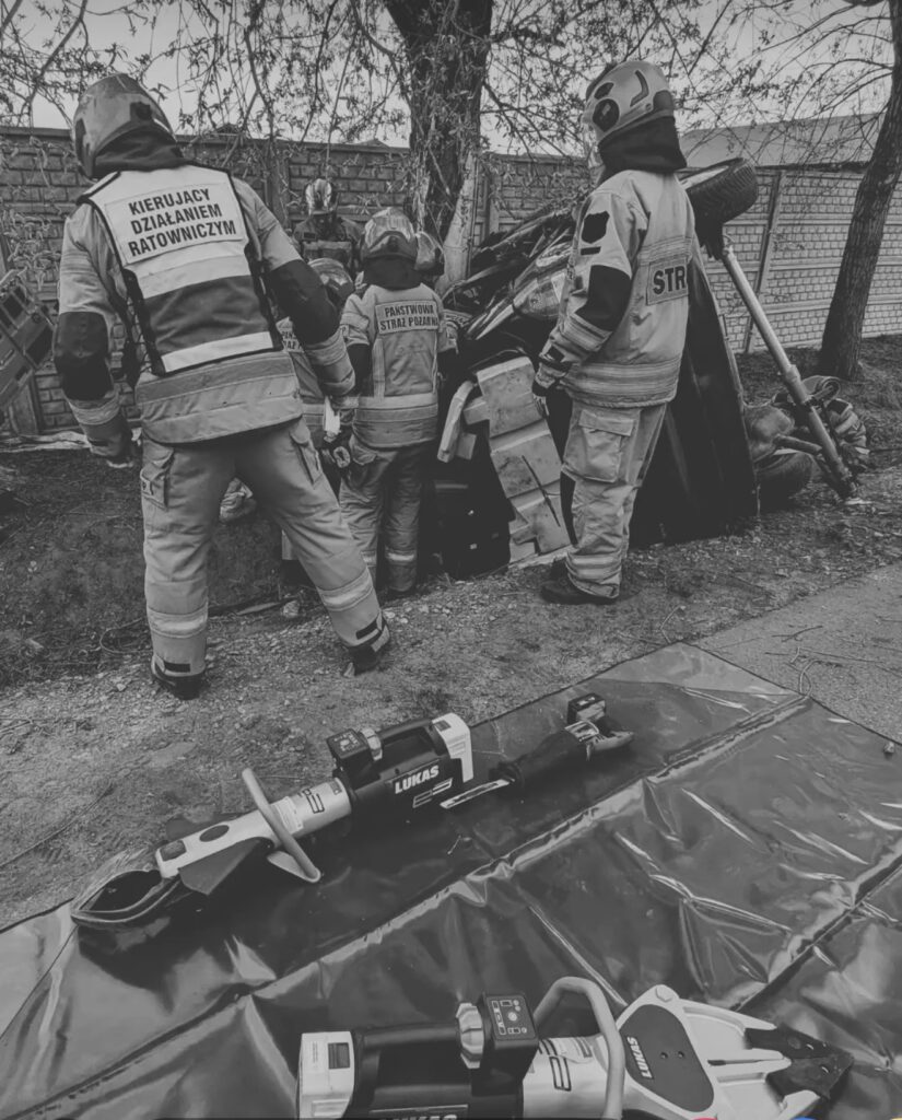 Czterech strażaków pracuje przy przewróconym pojeździe, otoczeni narzędziami ratunkowymi na ziemi.