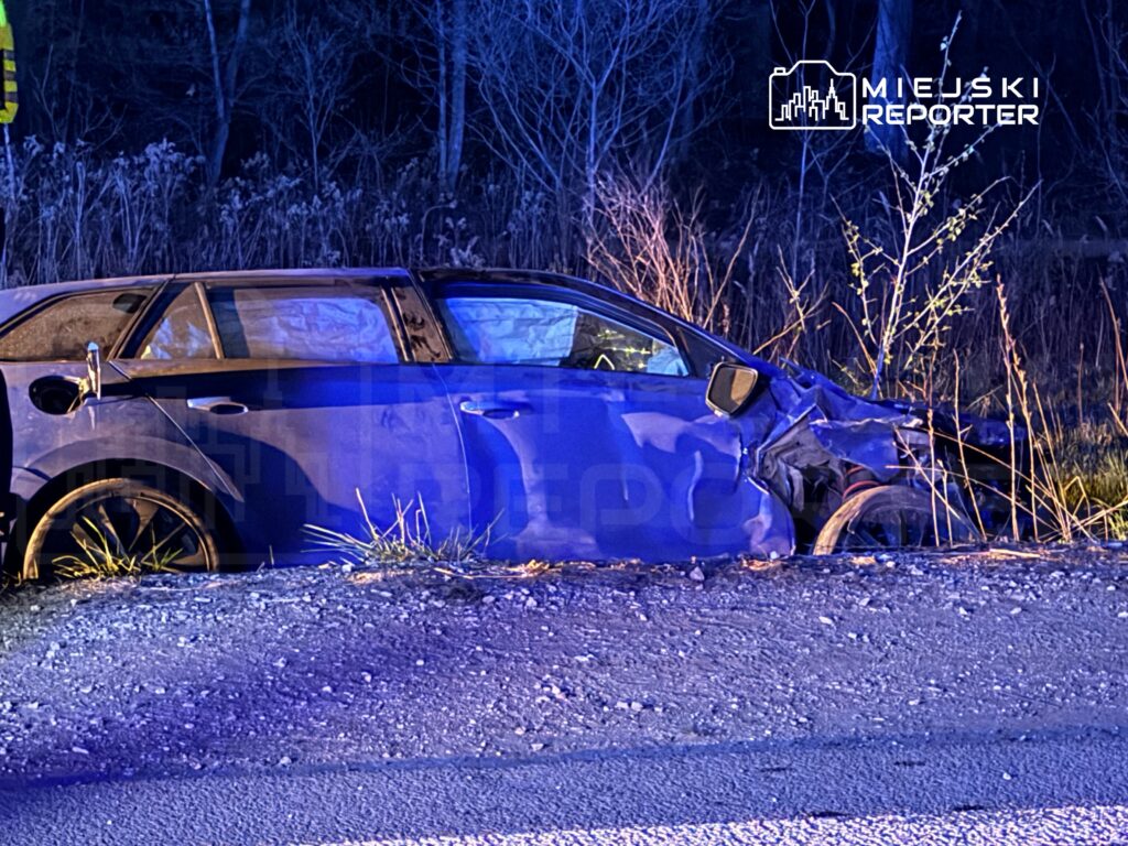 Uszkodzony niebieski samochód osobowy stoi na poboczu drogi, otoczony chwastami w nocy.
