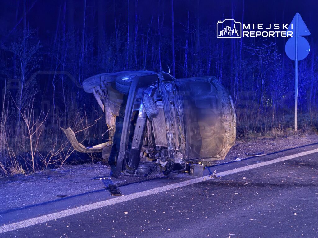 Przewrócony samochód leży na poboczu drogi, otoczony przez zarośla, w tle widać niebieskie światła.