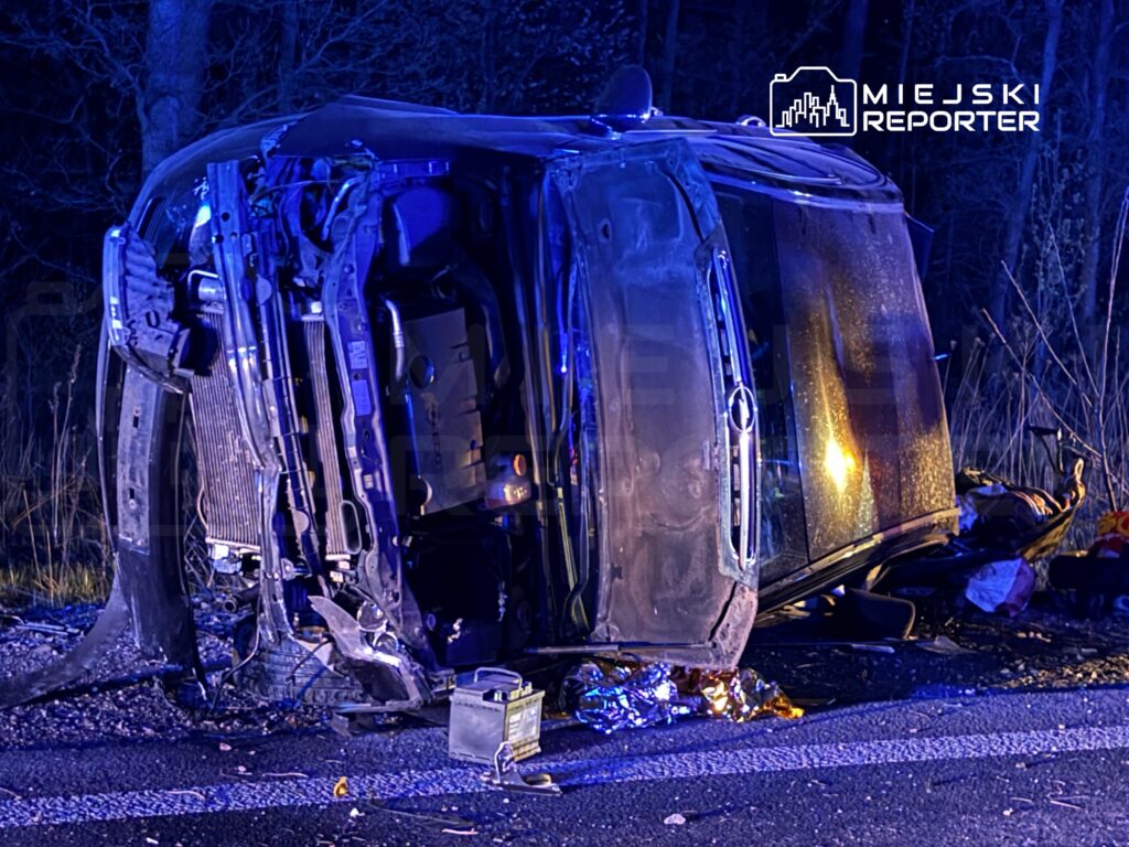 Przewrócony samochód osobowy leży na boku na poboczu drogi, w otoczeniu zarośli, pod niebieskim światłem.