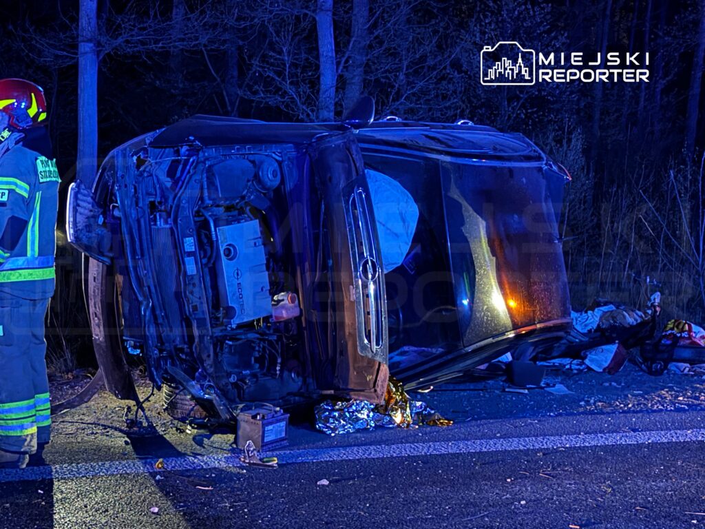Strażak w hełmie bada wypadek samochodowy, w którym pojazd leży na boku na poboczu drogi, otoczony ciemnym lasem.
