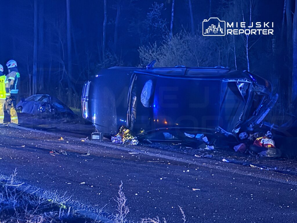 Ratownicy w odblaskowych strojach pracują przy przewróconym samochodzie na poboczu drogi w lesie.