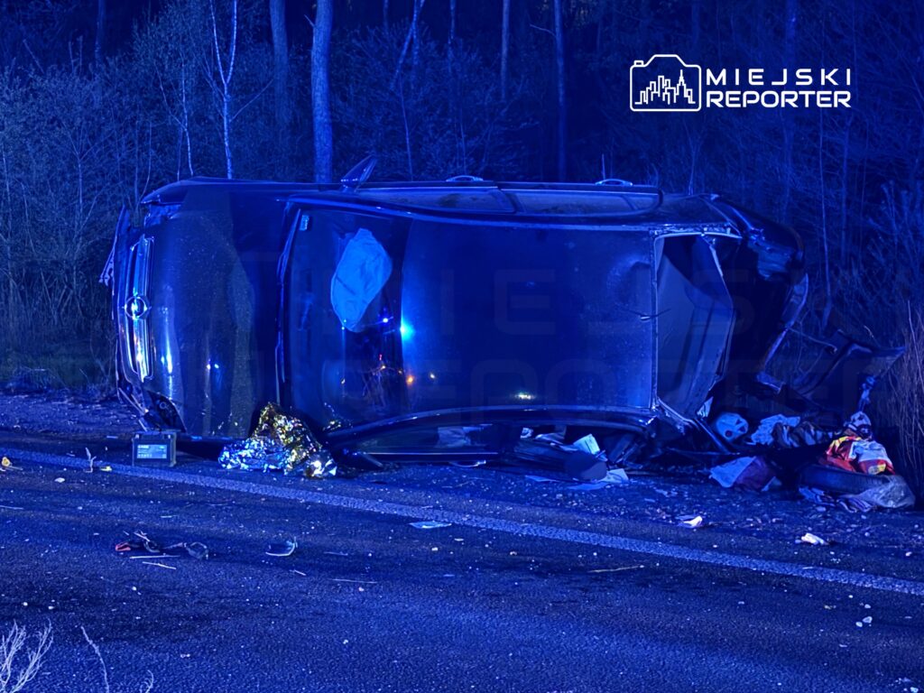 Przewrócony samochód osobowy leży na poboczu drogi, otoczony przez fragmenty szkła i odzież. W tle widoczne są drzewa, a scena oświetlona jest niebieskimi światłami.