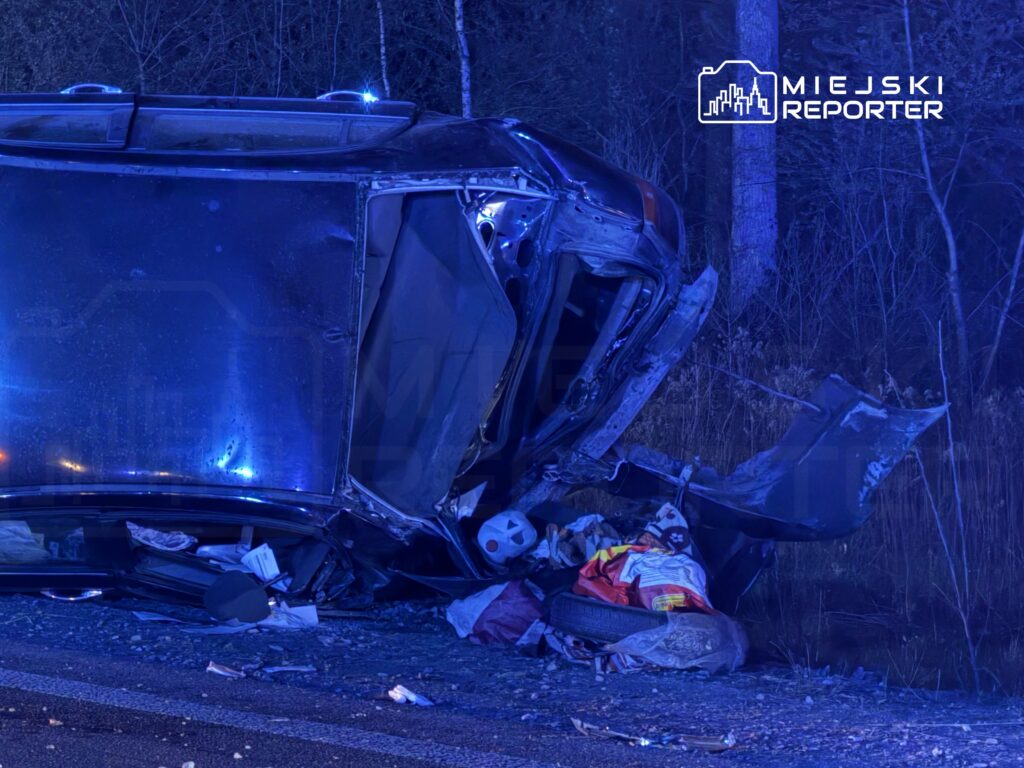 Przewrócony samochód osobowy leży na poboczu drogi, otoczony przez policyjne światła i zarośla.