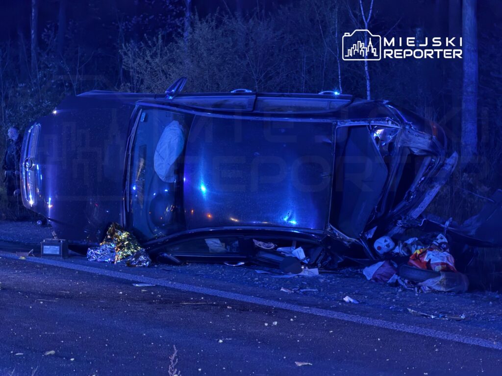 Samochód leży na dachu na poboczu drogi, w otoczeniu policyjnych świateł i porozrzucanych przedmiotów.